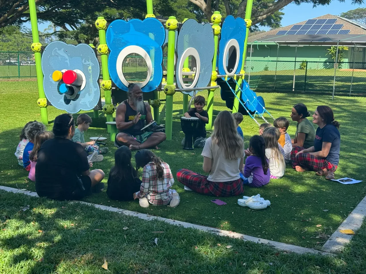 Keiki and adults seated on the grass with hand drums and shakers while a facilitator plays during an outdoor music session.