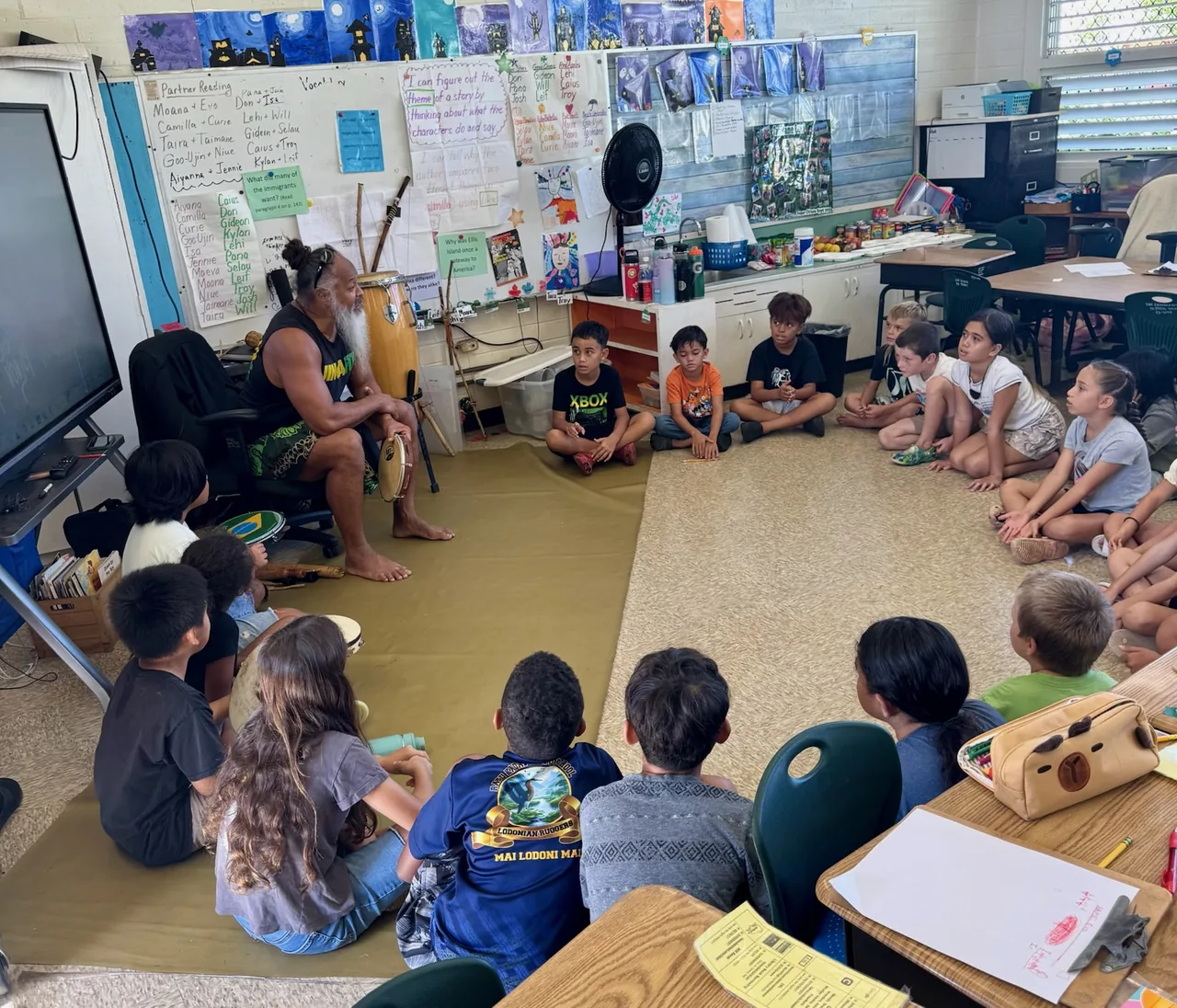 Elementary students seated on a classroom floor with hand percussion while a facilitator plays a drum.