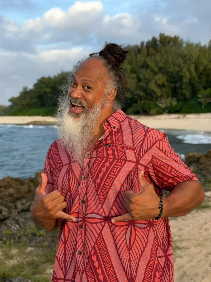 Branch Nautu smiling and throwing double shakas at the beach while wearing a red Polynesian-print aloha shirt.