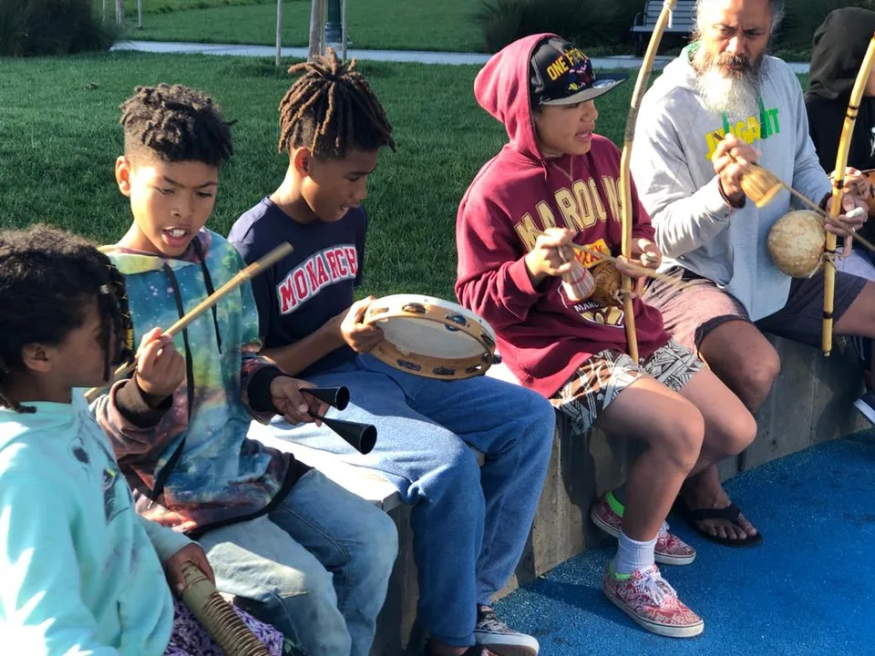 Keiki and an adult seated outdoors making music with handheld instruments.
