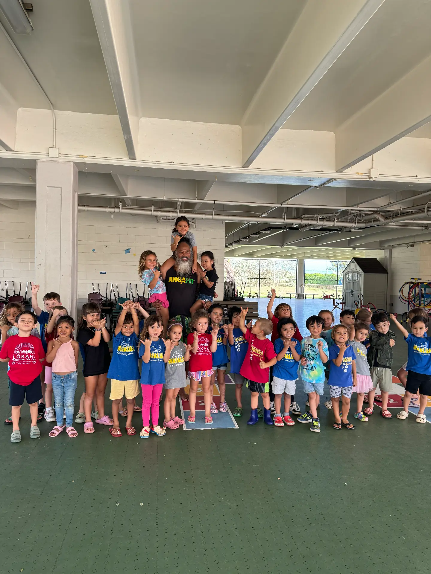 A large group of young children standing together with a facilitator in a covered school play area.
