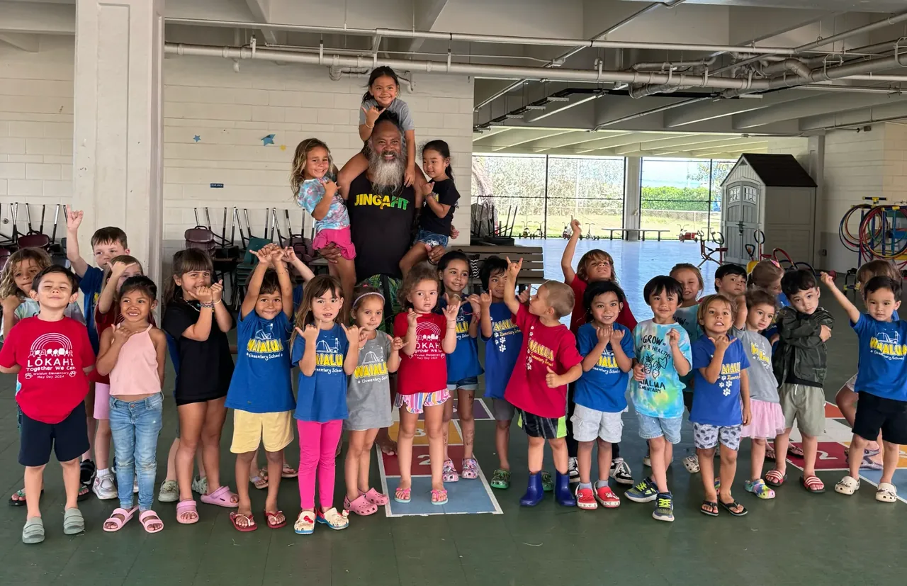 A large group of young children posing together with a facilitator in a covered school play area.