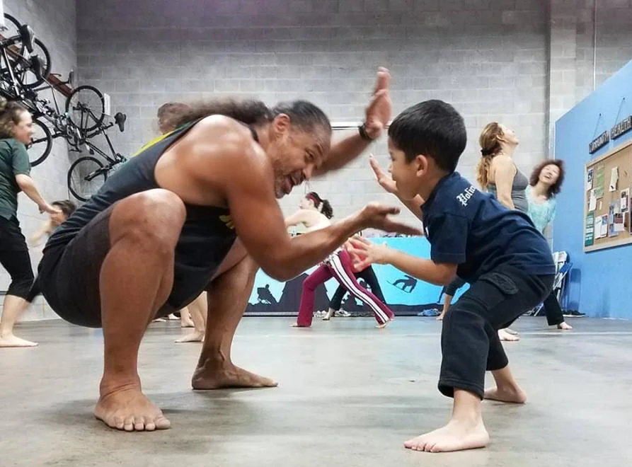 A facilitator and a young child face each other in low stances, mirroring each other's movement indoors.