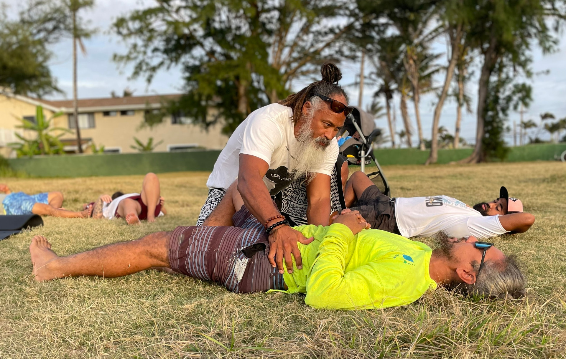 A CH³ facilitator supporting a participant through gentle restorative movement on the grass at a community gathering place in Koʻolauloa.